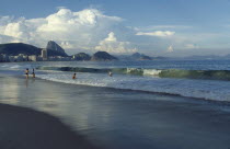 Beach with people playing in the surf.  City buildings and Sugar Loaf Mountain beyond. Brasil