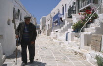 Folegandros.  Street scene with elderly man using stick to walk along pavement lined by white washed houses with steps leading from street to entrance.