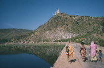 Women and children walking beside water reservoir tank twenty miles north of Udaipur.  Hindu shrine on hill top beyond.