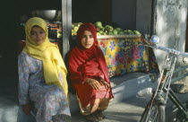 Two young girls sitting on step beside roadside fruit stall.