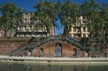 Toulouse.  Red brick steps leading down to the River Garonne from street level with facades of buildings partly seen through trees behind.  People sitting in the sun.