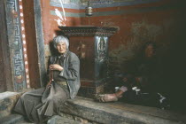 Man and elderly woman sitting on the floor beside a prayer wheel on a plinth