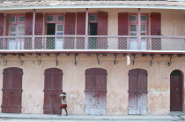 Street scene with woman walking past building with faded pink plaster walls and red doors.