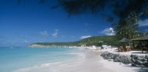 View along beach towards tree covered headland with people walking along shoreline and sitting under thatched sun umbrellas nearby.