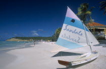 View along sandy beach lined with palms with boat in the foreground advertising Sandals hotel on its hull and sail.  Building rooftop part seen behind.