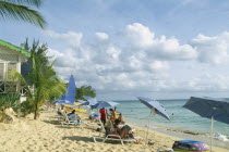 Mullins Bay.  People on loungers with blue sun umbrellas on sandy beach at the waters edge with palm tree and partly seen green and white building on wooden stilts.