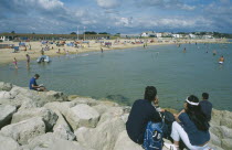 People sat on rocks looking across water and busy sandy beachfront