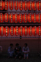 Teenage girls in yukata  summer kimono  sit beneath chochin lanterns each with the name of a donor to Narita san Temple during Gion Matsuri