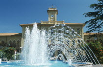 Fountains in town square with building with clock tower behind.