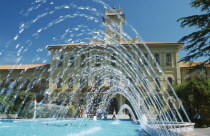 Fountains in town square with building with clock tower behind.