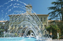 Fountains in town square with building with clock tower behind.