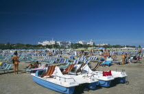 Busy beach scene with rows of striped sun loungers and pedalos in the foreground.