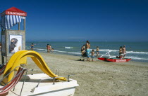 Adults and children on sandy beach with pedalo and lifeguard hut in foreground.