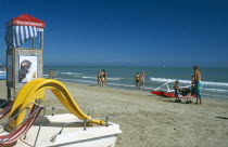 Adults and children on sandy beach with pedalo and lifeguard hut in foreground.