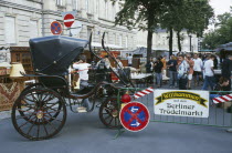 Antique carriage at entrance to flea market with people at stalls behind and market and road signs on crash barrier in foreground.