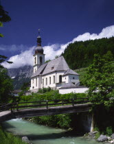 View over river and wooden bridge towards Ramsau Church and wooded hillside beyond.