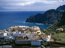 Spain,  Canary Islands, La Gomera Agulo, White yellow and orange painted houses with tiled rooftops, terraced hillside volcanic cliffs and sea beyond.