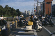 Commuters on scooters and bicycles waiting to cross road.