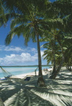 Akitua.  Sandy beach with people sunbathing on wooden sun loungers and hammock slung between palm trees in the foreground.