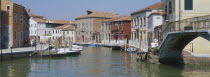 Murano Island.  View along canal lined by traditional houses with boats moored to posts at each side.