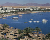 Naama Bay.  View over bay with jettys leading to tourist boats  encicled by sandy beach with thatched sun umbrellas and white sun loungers.  White painted buildings behind