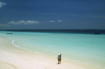 Nungwi.  View along beach with white sand and aquamarine sea  towards the skyline.  Couple walking along the shoreline in the foreground.