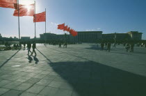 Great Hall of The People in silhouette with sun behind flying flags and people walking across square with long shadows