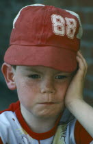 Young boy wearing red hat with face resting on his hand in sulky mood.