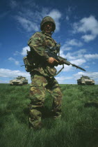 Soldier in camouflage standing in a field holding a gun with two Warrior tanks behind