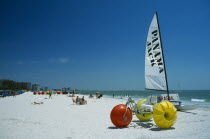 Hobbie cat and pedalo on stretch of white sand beach with few sunbathers.