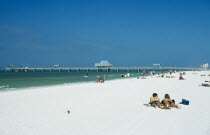 Sunbathers on white sand beach with pier beyond.