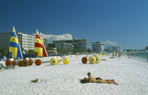 Sunbathers  Hobbie cats and pedalos on white sand beach with city buildings behind.