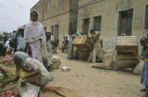 Market scene with women and children beside vegetable vendor with laden camels at side.