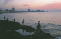 City skyline at sunset with fishermen beside the Malecon silhouetted against pink and purple sky reflected in the sea in the foreground.