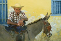 Portrait of elderly Cuban man smoking a cigar and leaning on saddle of blinkered donkey.