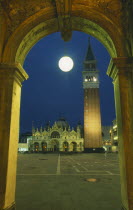 Piazza di San Marco.  Part view of square with Basilica di San Marco and the Campanile framed by stone archway with central circular lamp illuminated at night.St Mark