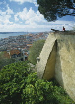 Visitor sitting on fortified walls beside cannon of Castelo de Sao Jorge  with rooftop view across city.