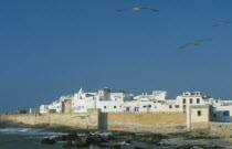 Fortified coastal town with waves breaking against rocks in the foreground.