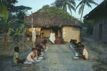 Outdoor informal school. Pupils seated in two lines facing each other with teacher at head.