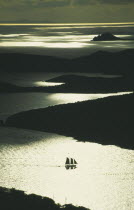 Silhouette of two masted sailing ship with three sails up amongst the bays of the coastline