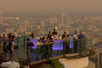 Sorocco Restaurant. People standing next to a lit up bar and looking over balcony with the Bangkok Skyline in the background.  Taken from the State Tower the 2nd tallest building in Bangkok