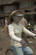 Woman wearing a conical hat with a scarfe covering her mouth riding a bycicle