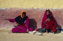 Settled Bedouin woman spinning wool with older woman and child sitting beside her against faded ochre and terracotta painted wall.