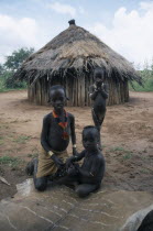 Hamer children sitting on animal skin outside thatched hut.