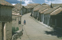 Town in the Altiplano or Bolivian Plateau.  Woman carrying two buckets walking along narrow paved street between houses with overhanging tiled rooftops.