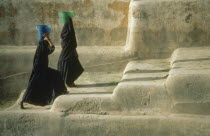 Two veiled women fetching water carrying buckets on their heads. Walking up steps from village cistern.