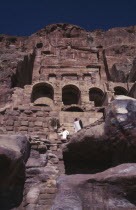 View looking up at historical tombs carved in sandstone cliffs with two men dressed in white looking downwards