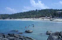 View from rocky outcrop over bay toward beach surrounded by palms with group of bathers in the sea