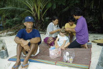 Young Western girl on sandy beach having her hair braided by three Thai women with man sitting smoking in front and palm trees behind.