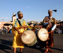 Two men playing the drums  wearing costumes and hats with tassles  standing in the street Marrakesh